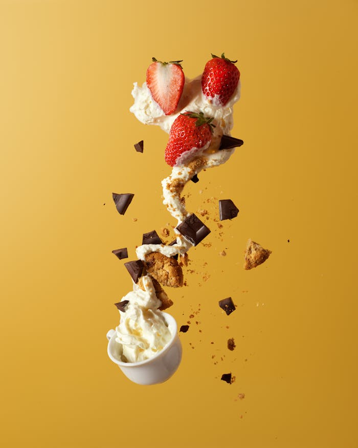 menu-02 Delicious strawberries and cream with chocolate and cookie pieces, captured in mid-air against a vibrant yellow background.